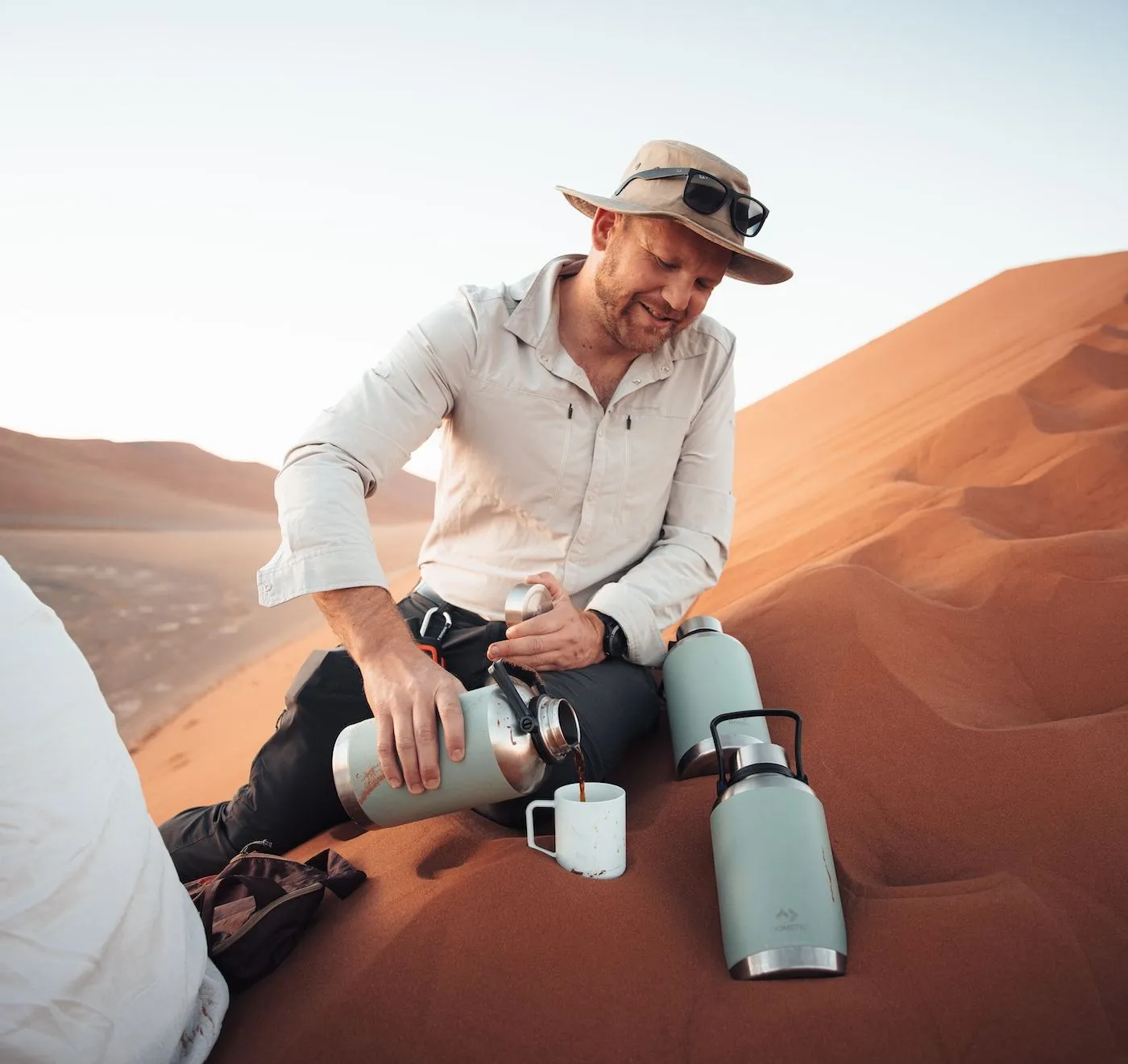 Mann mit Hut gießt Kaffee aus der Thermoskanne auf der Düne