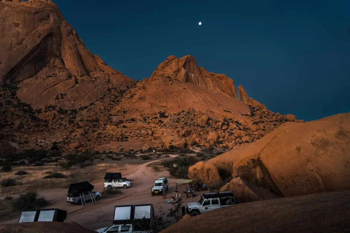 Fahrzeuge am Fuß der Spitzkoppe, der Mond steht schon am Himmel